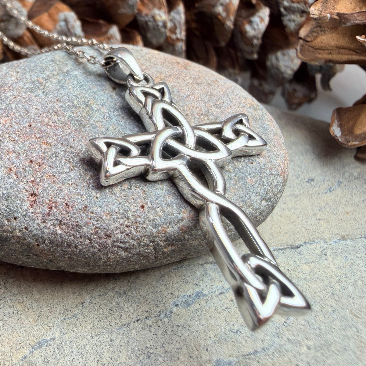 A silver Celtic knot cross pendant with a center trinity knot on a matching chain, displayed on a smooth gray stone surrounded by pinecones.