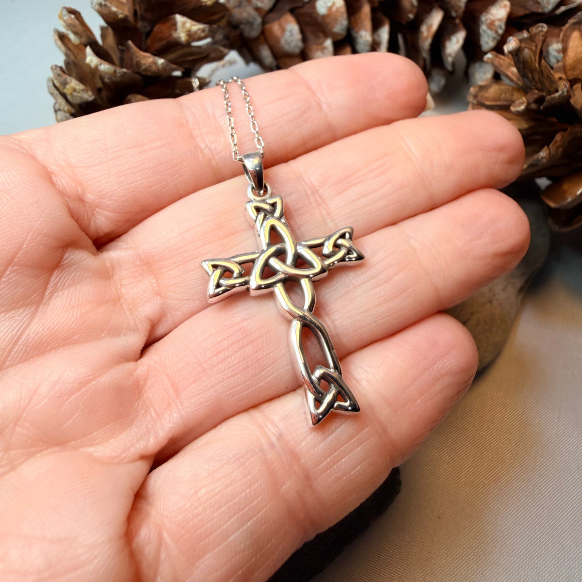 A silver Celtic knot cross pendant necklace resting on a textured gray stone, with a smooth beige stone in the background.