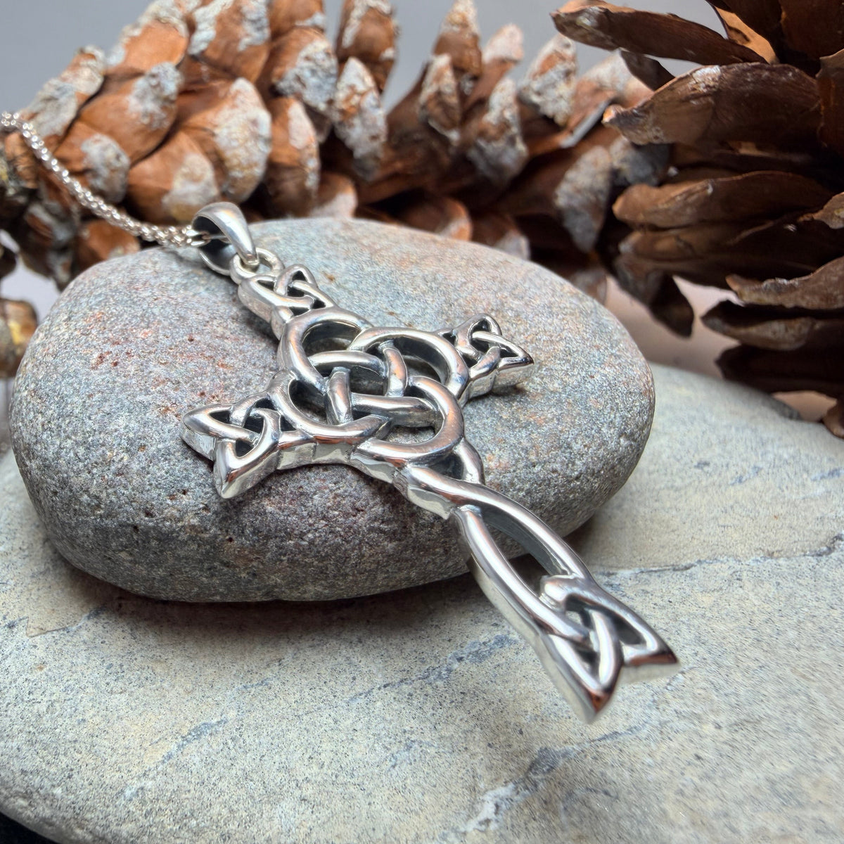 A silver Celtic knot cross pendant with  a center dara knot on a chain, displayed on a smooth gray stone with pinecones in the background.