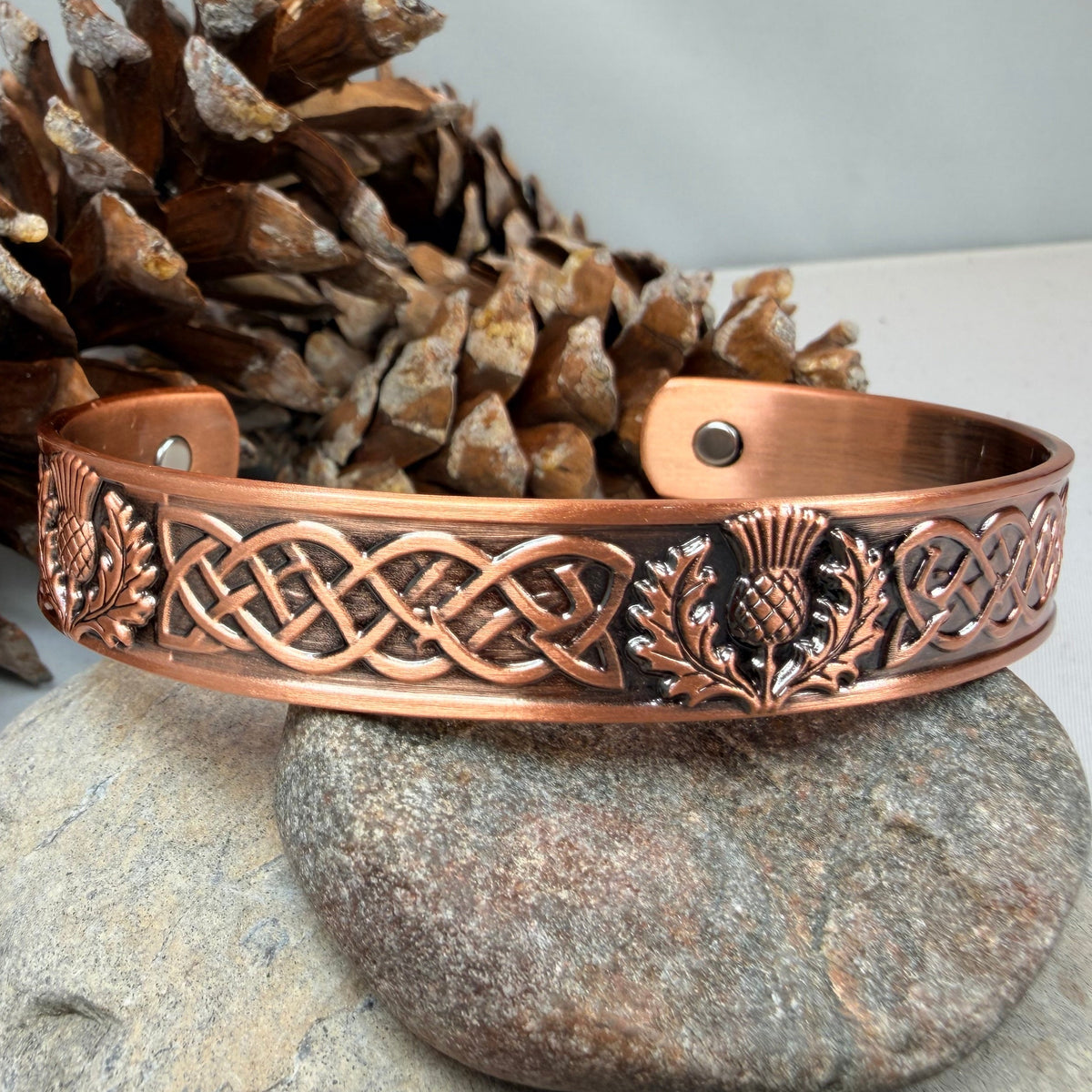 A copper-colored metal cuff bracelet with Celtic knotwork and thistle motifs, displayed on a stone with pinecones in the background.