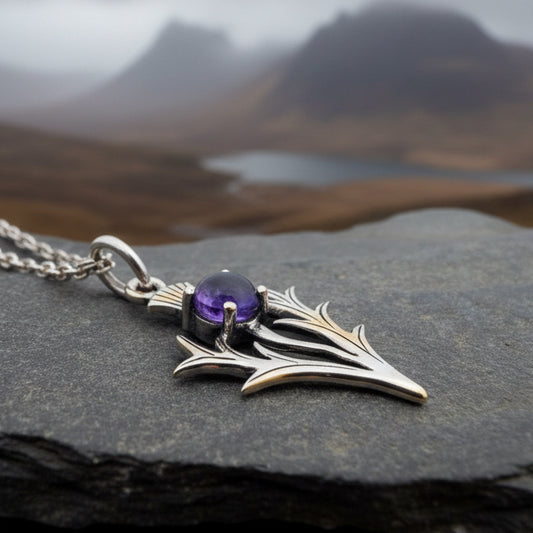A silver-toned pendant necklace with a thistle design, featuring a purple amethyst gemstone, displayed on a dark gray stone surface.