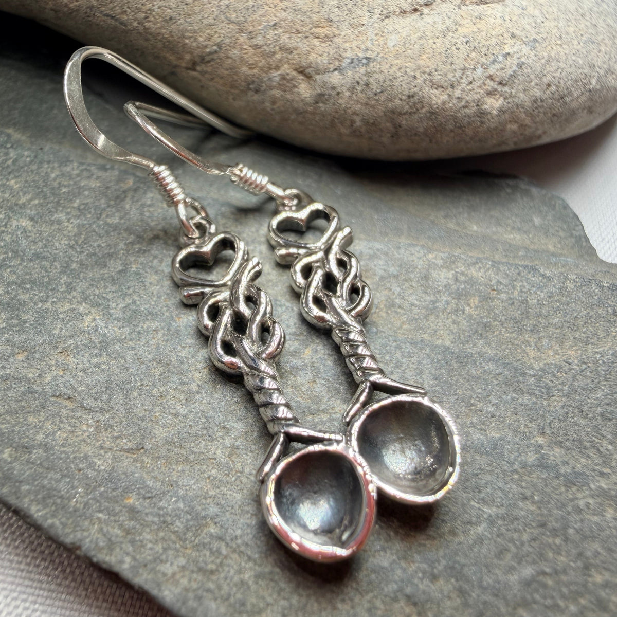 A pair of silver-toned dangle earrings with ornate filigree details and oval clear stone drops, displayed on a gray stone surface with a larger stone in the background.