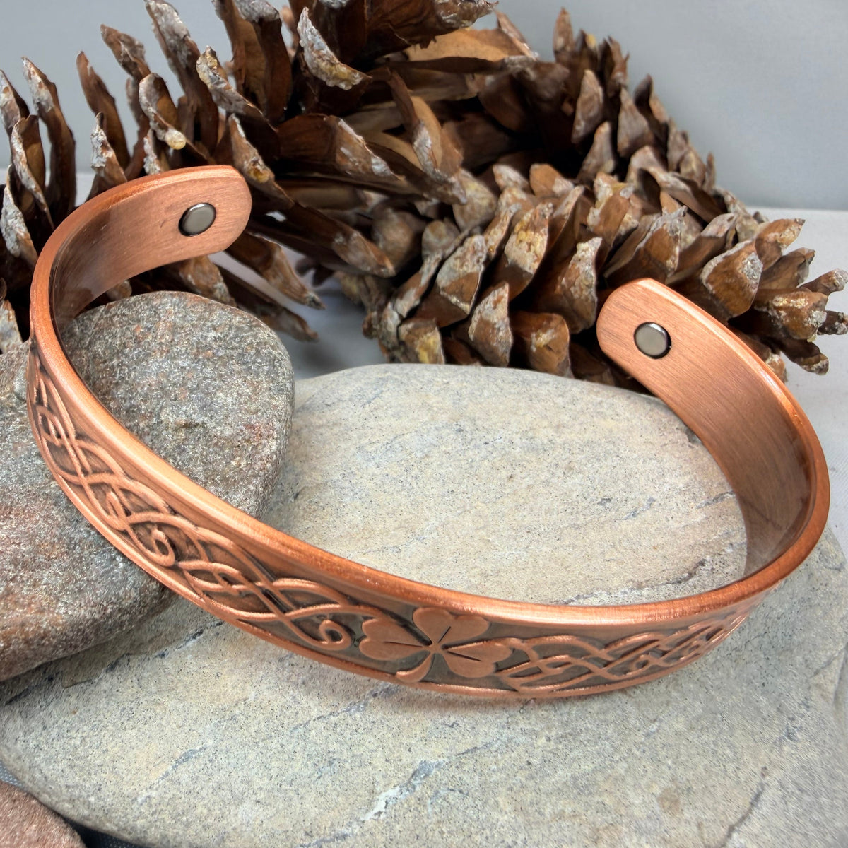 A copper cuff bracelet with engraved floral and geometric patterns, displayed on a smooth gray stone with pinecones and other rocks in the background.