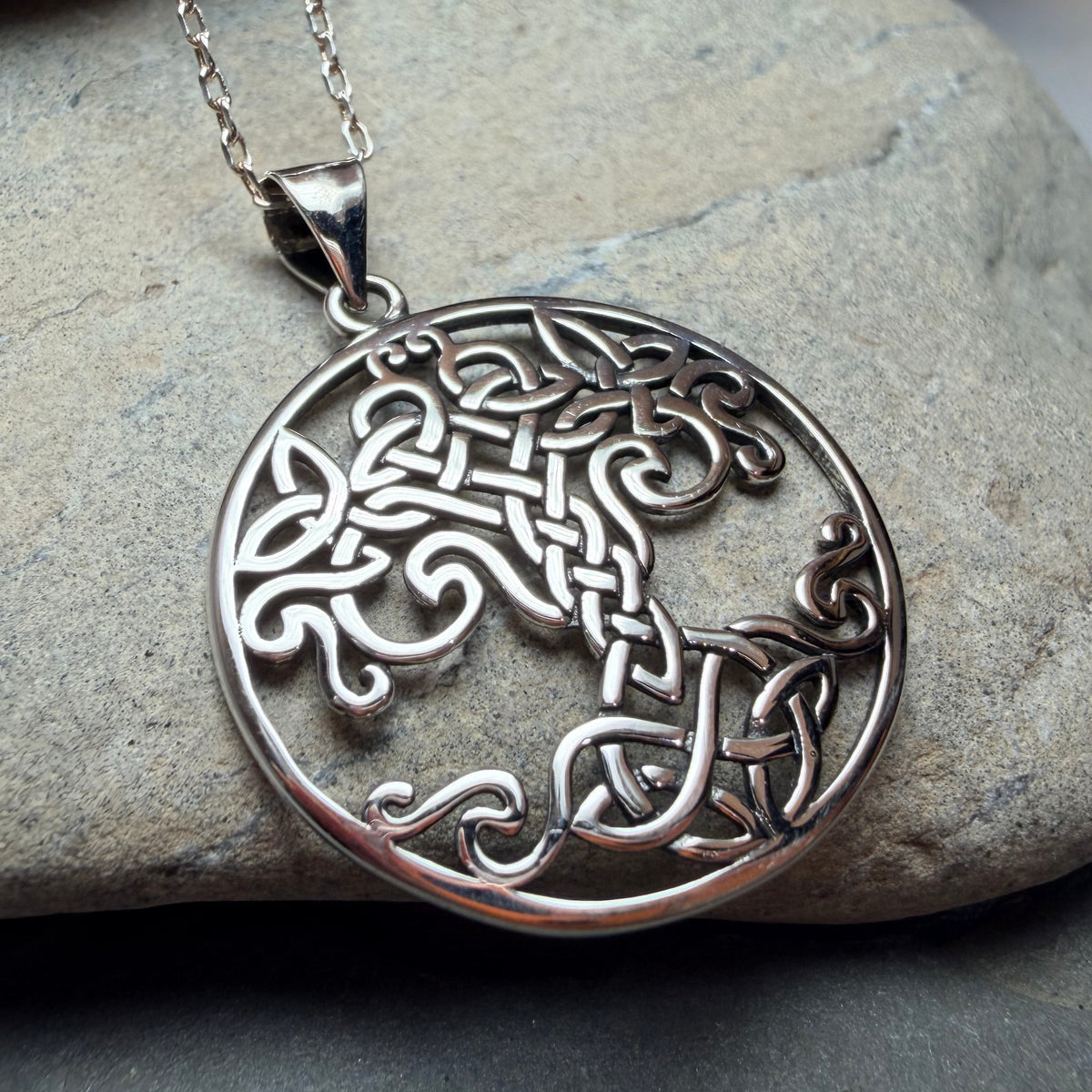 A silver Celtic tree knotwork pendant necklace resting on a smooth gray stone, with a matching silver chain extending upwards against a soft gray and white background.