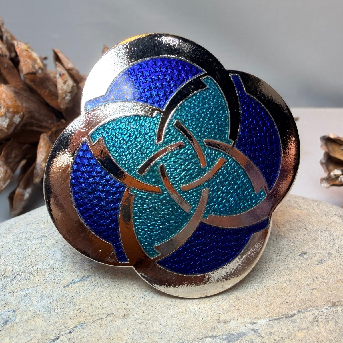 A round enamel pin featuring a Celtic triquetra knot design in turquoise and deep blue with metallic copper borders, displayed on a light stone with pinecones in the background.