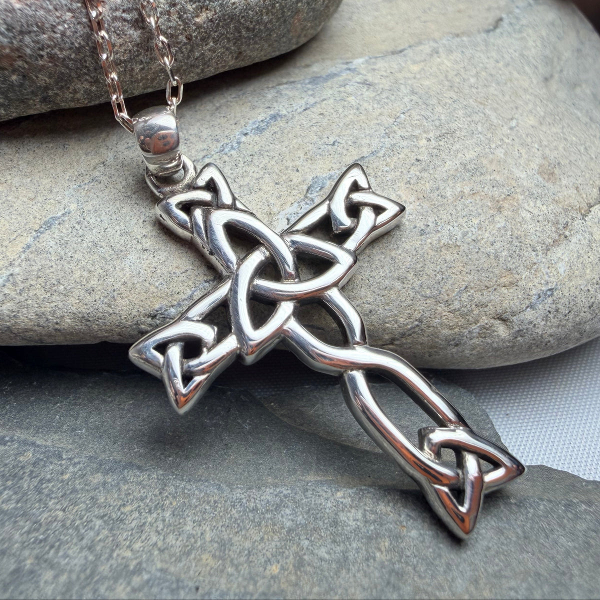 A silver Celtic cross pendant necklace resting on smooth gray stones, with a pinecone in the background.