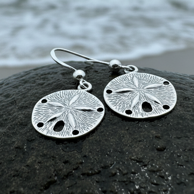 A pair of silver-toned dangle earrings shaped like sea urchins or sand dollars, resting on a wet dark rock with blurred ocean water in the background.