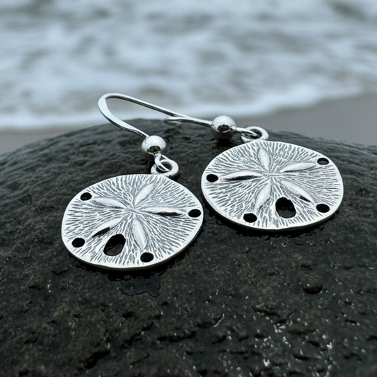 A pair of silver-toned dangle earrings shaped like sea urchins or sand dollars, resting on a wet dark rock with blurred ocean water in the background.