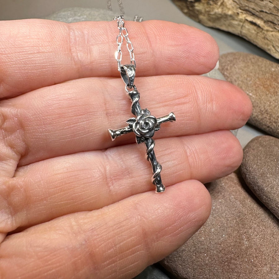 Silver cross necklace held between fingers with pebbles in the background