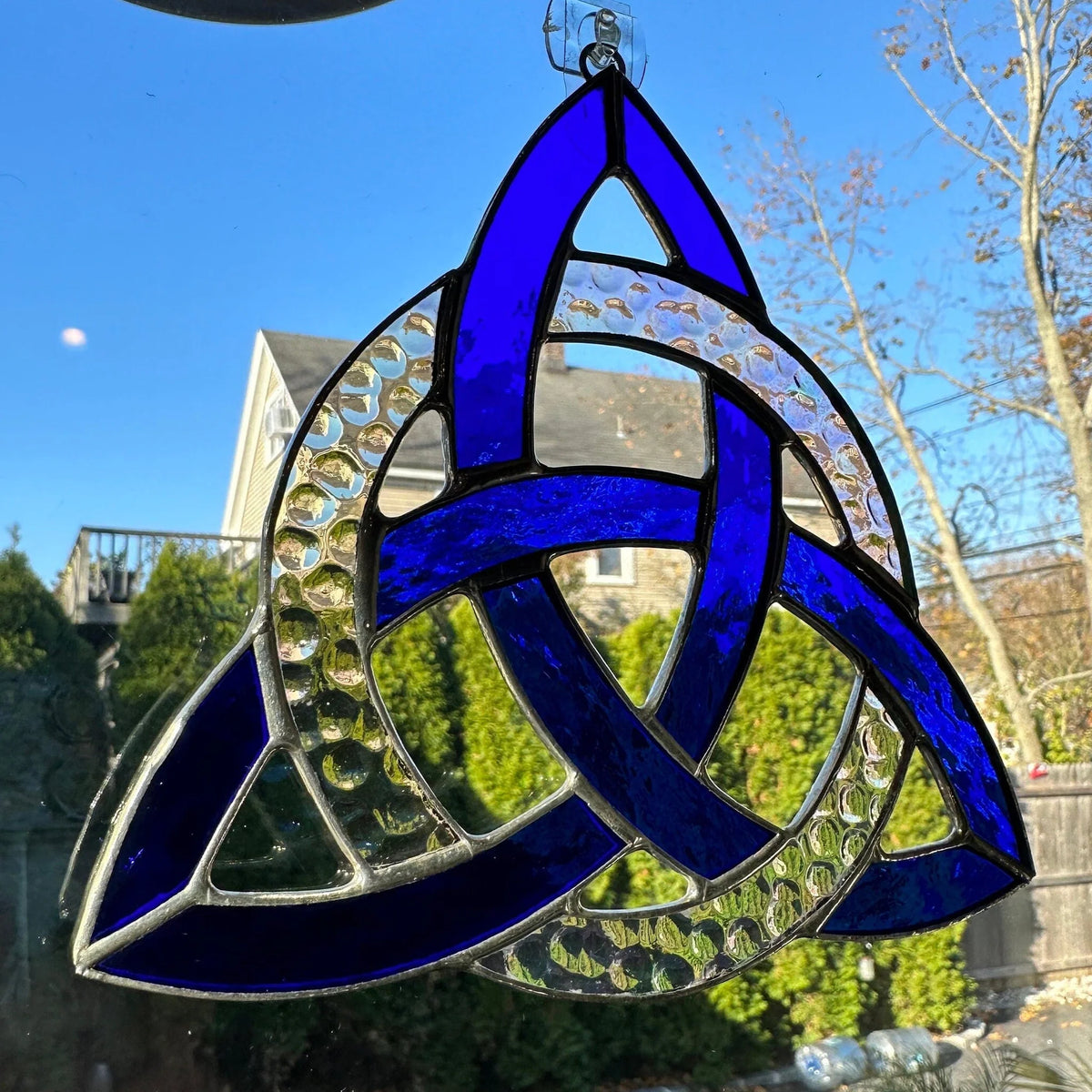 Blue stained glass Trinity knot hanging outdoors with a clear blue sky and trees in the background.